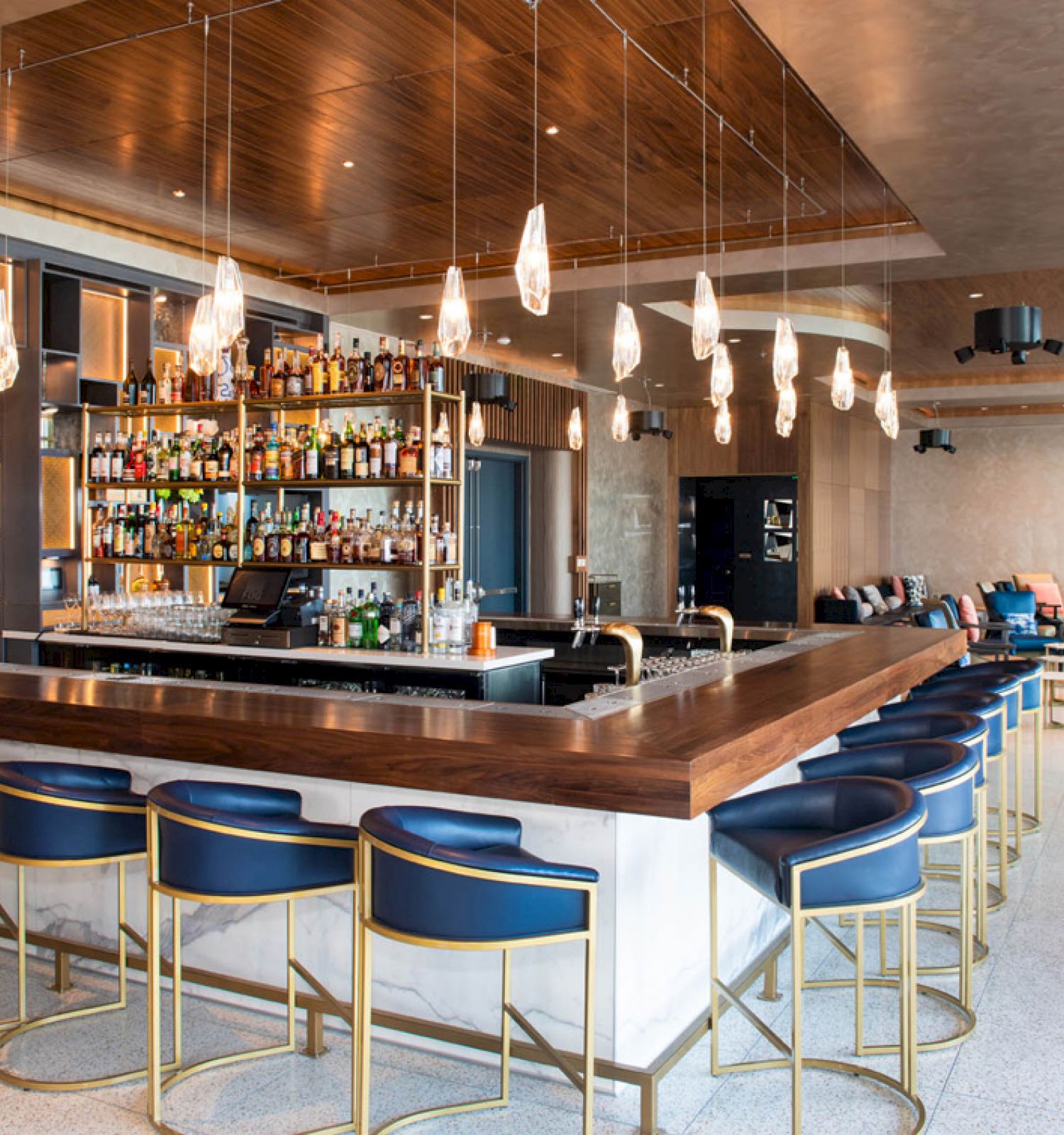 The image shows a modern bar with a wooden counter, blue bar stools, hanging lights, and a well-stocked liquor shelf. There’s seating in the background.