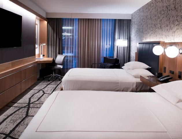 The image shows a modern hotel room with two neatly made beds, a flat-screen TV, desk, chair, lamps, and stylish decor.
