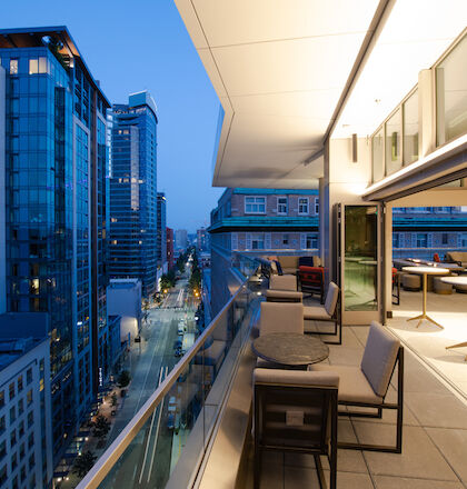 A modern cityscape featuring a high-rise building with a balcony, furnished with tables and chairs, overlooking a street at dusk, with the city lights on.
