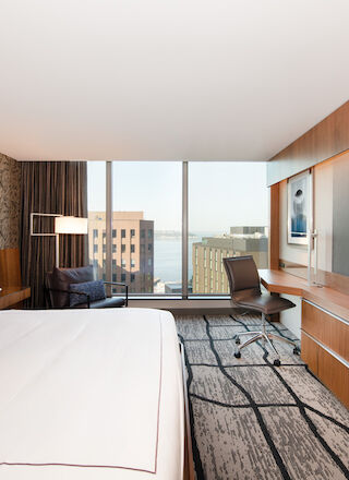 A modern hotel room with a large bed, a desk with a chair, a sofa, a flat-screen TV, and a city view through the window.