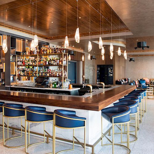 A modern bar with a wooden countertop, blue bar stools, a well-stocked liquor shelf, pendant lights, and a seating area with a contemporary design.