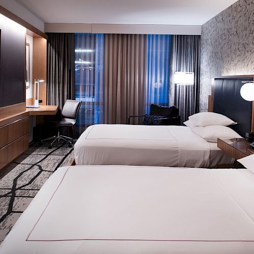 A modern hotel room with two neatly made beds, a wall-mounted TV, a desk with a chair, and stylish lighting and decor, is shown in this image.