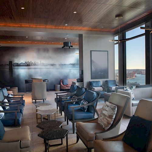 A modern lounge area with multiple chairs, tables, a wall mural, and large windows offering a scenic view of a Ferris wheel and water.