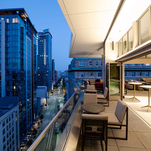 A modern balcony with seating overlooks a cityscape at dusk. The interior has additional tables and chairs, with glass windows open to the outside.