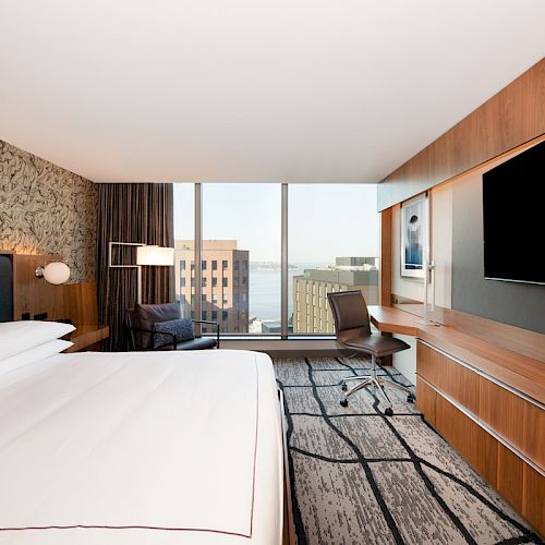A modern hotel room with a large bed, desk, chair, TV, and expansive windows offering a city view with a glimpse of water.