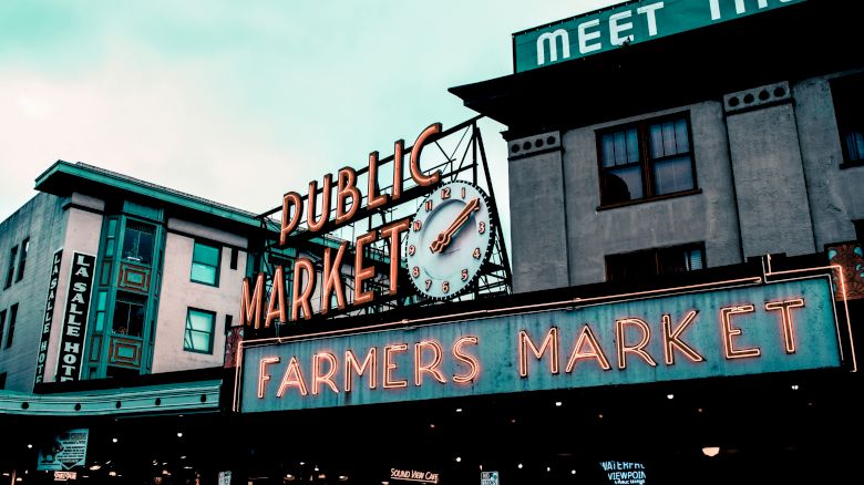 The image shows a public market building with neon signs reading "Public Market" and "Farmers Market" under a clock. Other buildings are visible too.