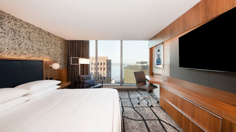A modern hotel room with a large bed, a wall-mounted TV, a desk, and a view of city buildings through a large window ends the sentence.