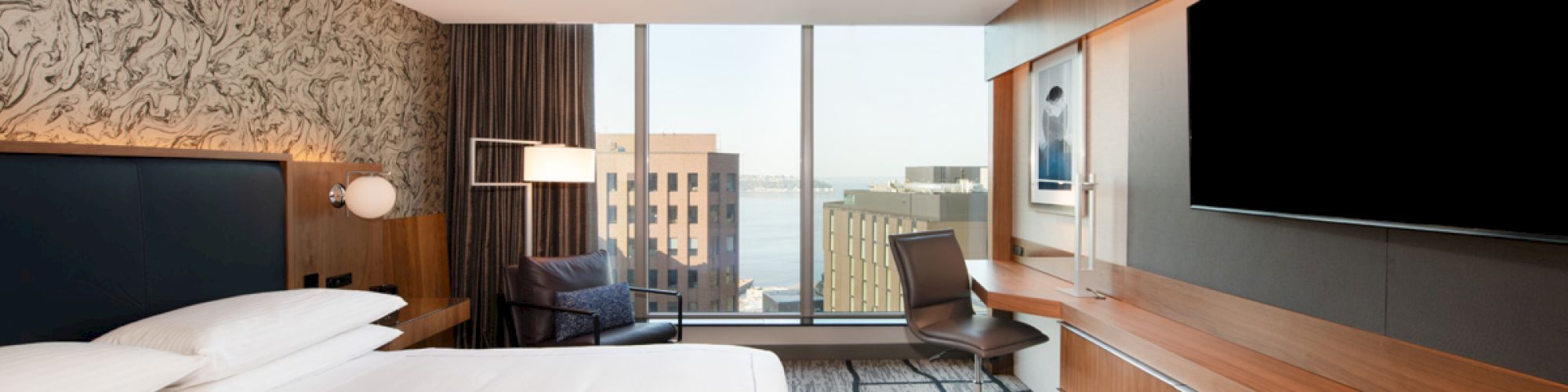 The image shows a modern hotel room with a large bed, a desk with a chair, a TV, and a window with a city view.