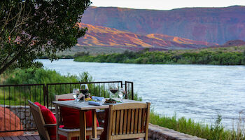 A table with food and wine is set on a deck overlooking a river and red rock hills, with trees around.