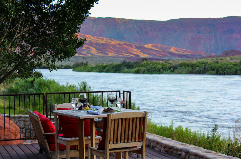 A table with food and wine is set on a deck overlooking a river and red rock hills, with trees around.