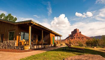 A rustic cabin with wooden details sits by a red rock formation under a blue sky with clouds, surrounded by grass and nature.