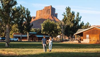 A couple walks hand in hand toward a cabin, with a large rock formation and trees in the background.