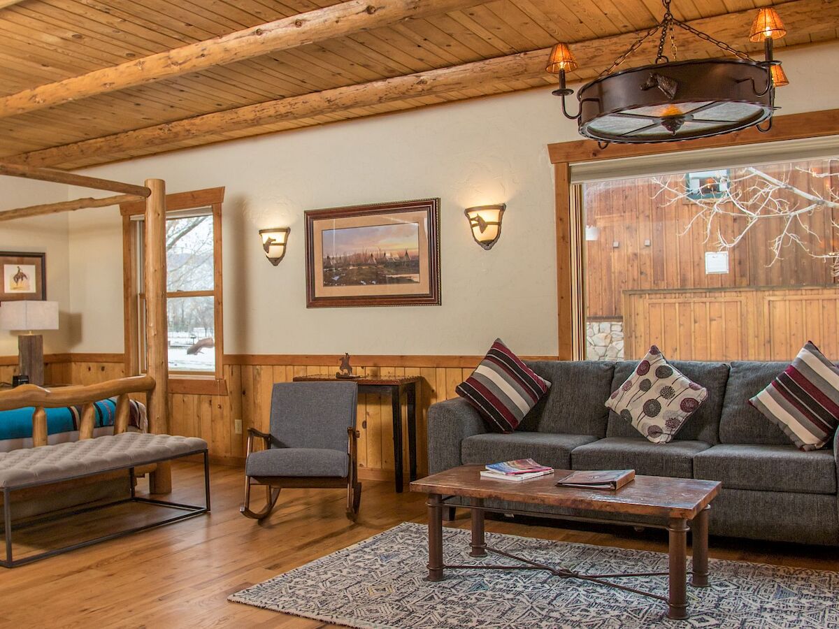 The image shows a cozy wooden living room with a sofa, armchair, coffee table, bed, framed pictures, and lamps, creating a warm, inviting atmosphere.