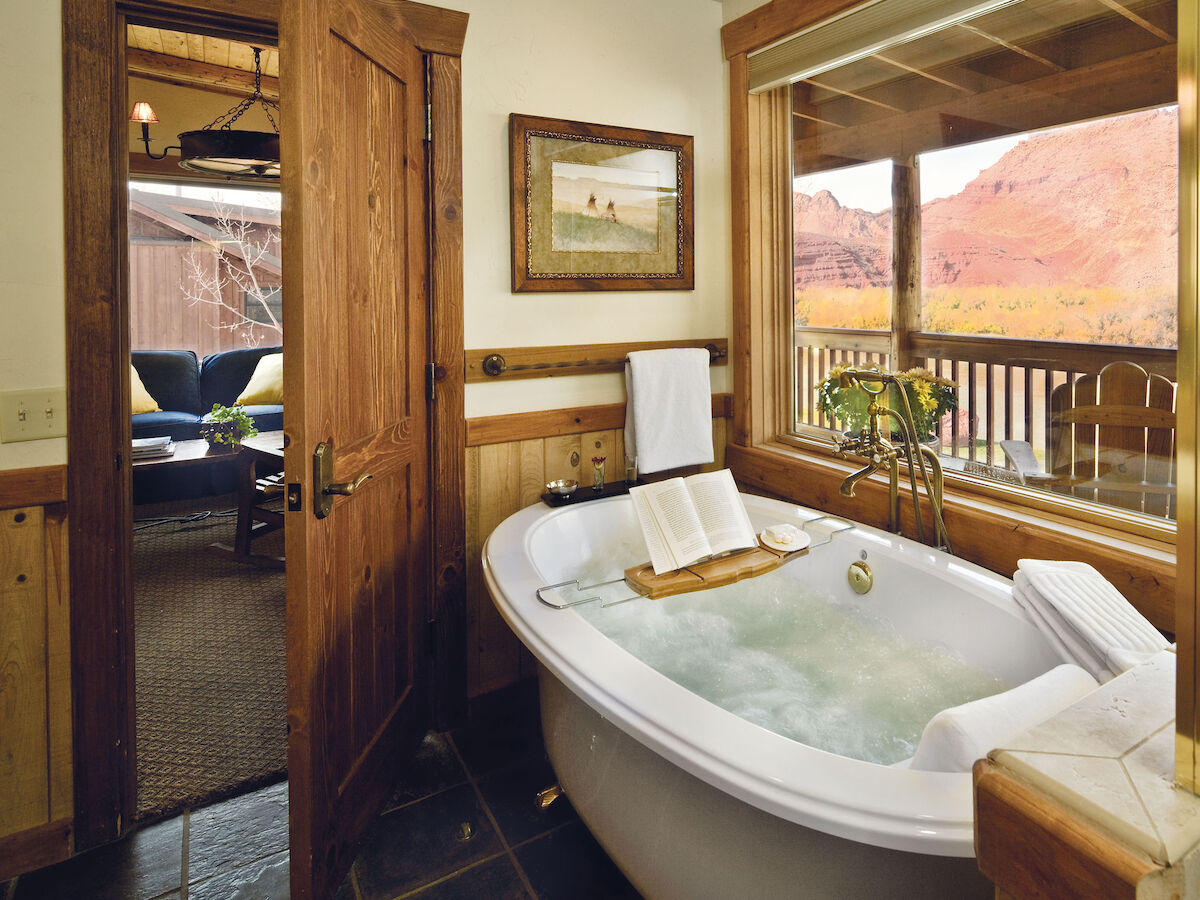 A cozy bathroom with a bathtub, book on tray, large window with scenic view, wooden door, and a glimpse of a seating area outside.