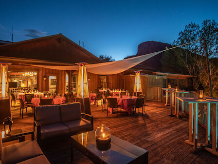 A cozy outdoor dining area with tables and chairs, lit by hanging lights and candles, surrounded by trees and a scenic view.