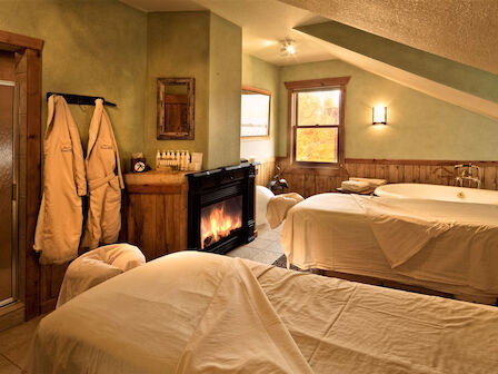 A cozy spa room with two massage tables, a bathtub, fireplace, robes, and ambient lighting, creating a relaxing and warm atmosphere.