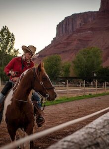 A person in a cowboy hat riding a horse in a fenced area, with tall red rock formations and trees in the background.